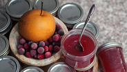 Pear and cranberries beside open jar of red jam with spoon. Multiple sealed jars in the background on a speckled surface.