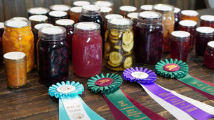 Jars of preserved foods are displayed on a wooden table with award ribbons in blue, purple, and teal labeled "Best of Class."