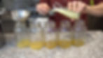 Hands pouring yellow liquid into mason jars using a glass pitcher and measuring cup on a speckled countertop, kitchen in background.