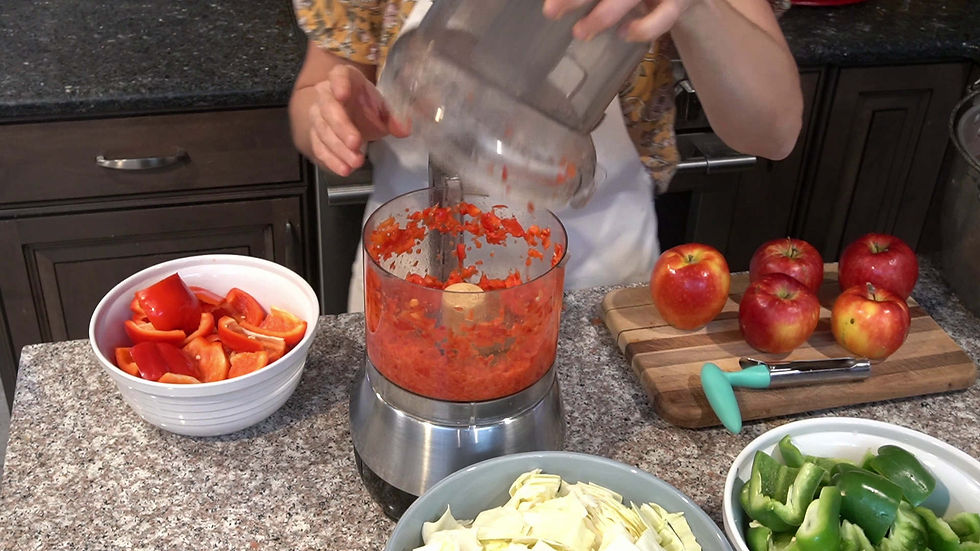 A person processes red bell peppers in a food processor. Nearby are apples on a cutting board and bowls of vegetables in a kitchen setting.