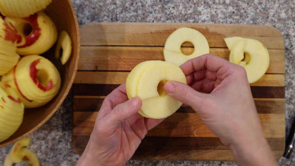 Hands holding sliced apple rings on a wooden cutting board. More peeled apple slices in a bowl nearby. Kitchen countertop background.