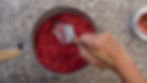 Hand using a spatula to press red raspberries through a metal sieve over a speckled countertop; nearby jar lid visible.