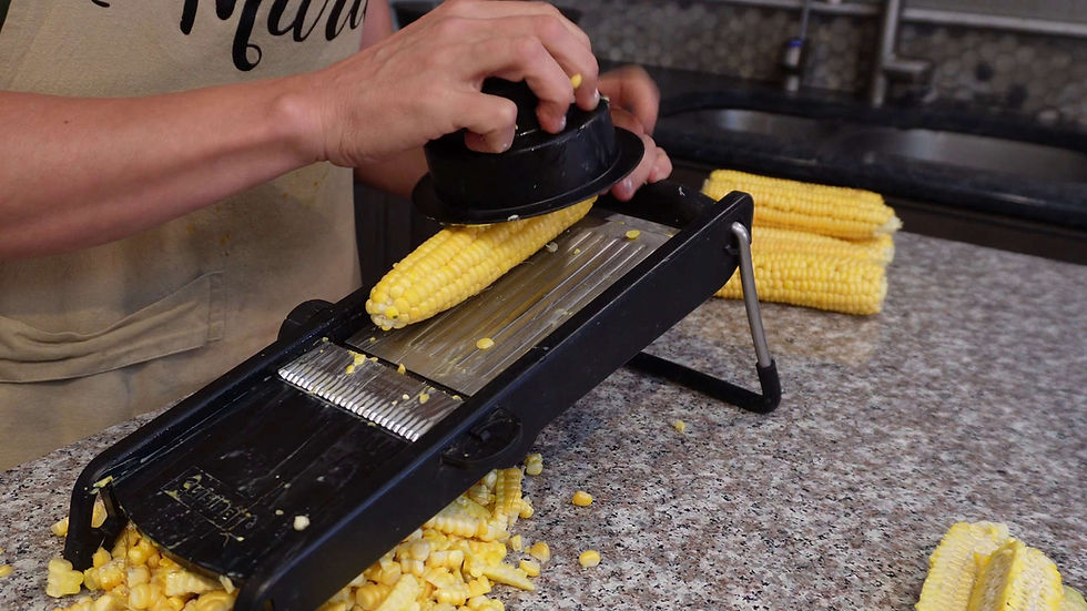 Person using a black corn stripper on a yellow cob over a granite countertop, with kernels scattered and bundled cobs in the background.