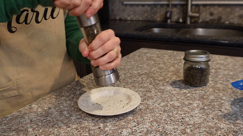 Hands using a pepper grinder over a white plate on a granite countertop. Clear jar with peppercorns nearby. Person wears a green shirt and apron.