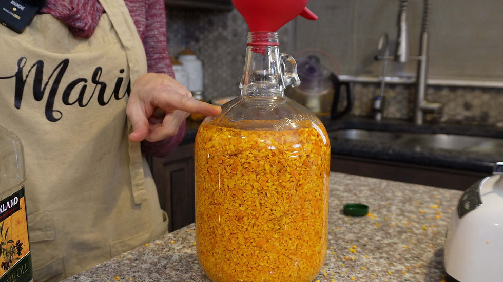 Person points at a glass jug filled with chopped orange material on a kitchen counter. They wear an apron with "Mari" text. A red funnel is on the jug.