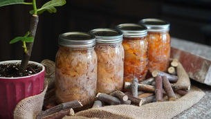 Potted plant in a pink pot and four mason jars with canned pork on burlap, surrounded by small wooden sticks on a stone surface.