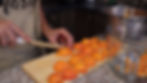 Person chopping apricots on a wooden board in a kitchen. Wearing a beige apron, next to glass and metal bowls. Cozy and focused atmosphere.