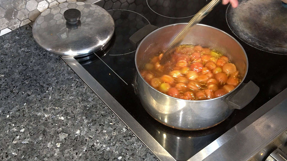 Pot of cherry tomatoes simmering on a stovetop, stirred by a hand. Granite countertop and silver lid nearby. Cozy kitchen vibe.