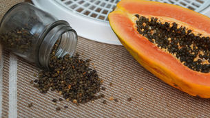 A halved papaya with black seeds is next to a jar spilling seeds onto a striped brown surface. The setting appears kitchen-like.