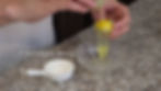 Hands separate an egg over a glass bowl on a speckled countertop, with the text "1 teaspoon Water" and a white measuring spoon nearby.