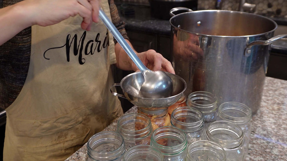 Person in an apron marked "Marie" funnels liquid into jars from a large pot on a kitchen counter. Glass jars are arranged nearby.