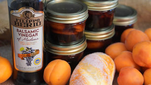 Balsamic vinegar bottle, jars of preserves, fresh apricots, and a sugar-coated pastry on a burlap surface. Warm, rustic feel.
