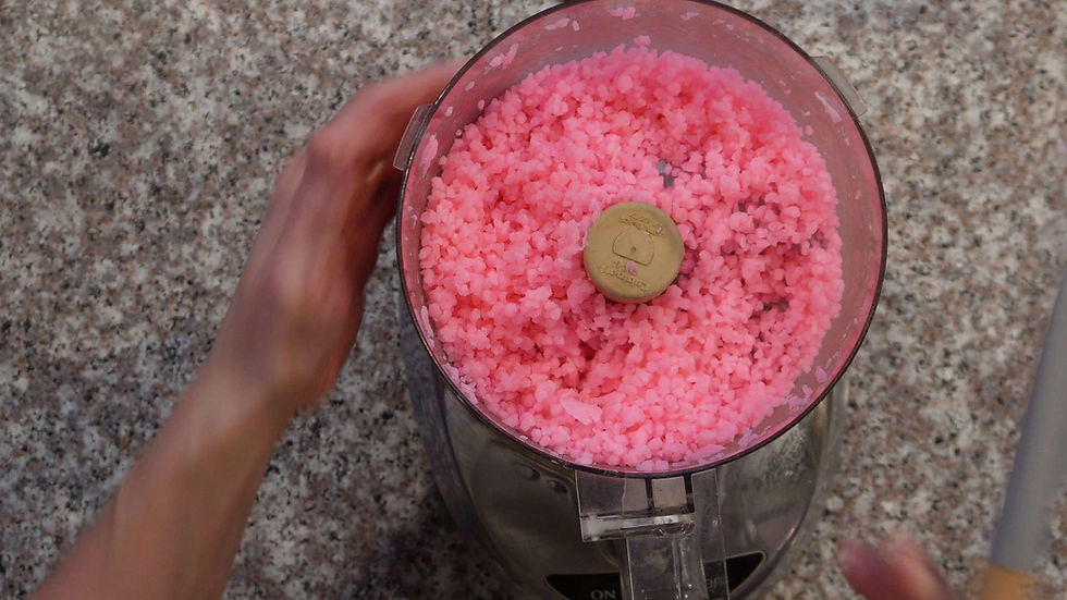 Hands near a food processor filled with pink crushed ice on a speckled countertop. The mood is calm and focused.