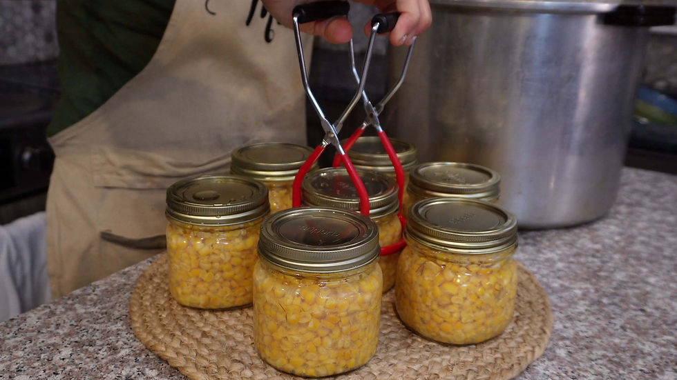 Hand placing sealed jars of corn onto a woven mat. A pot and apron-clad person are in the background. Jars are golden and gleaming.