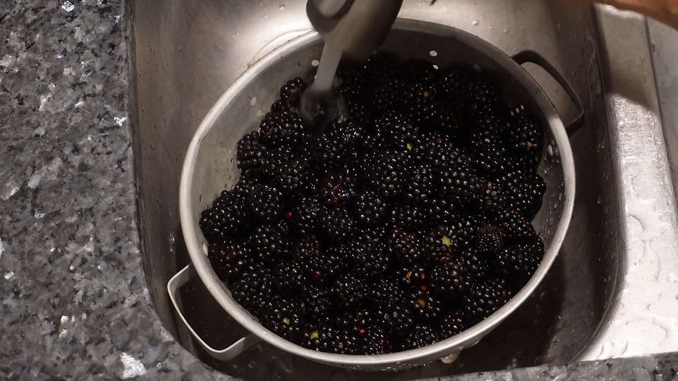 Blackberries being rinsed under running water in a metal colander, set in a stainless steel sink. The scene conveys freshness.