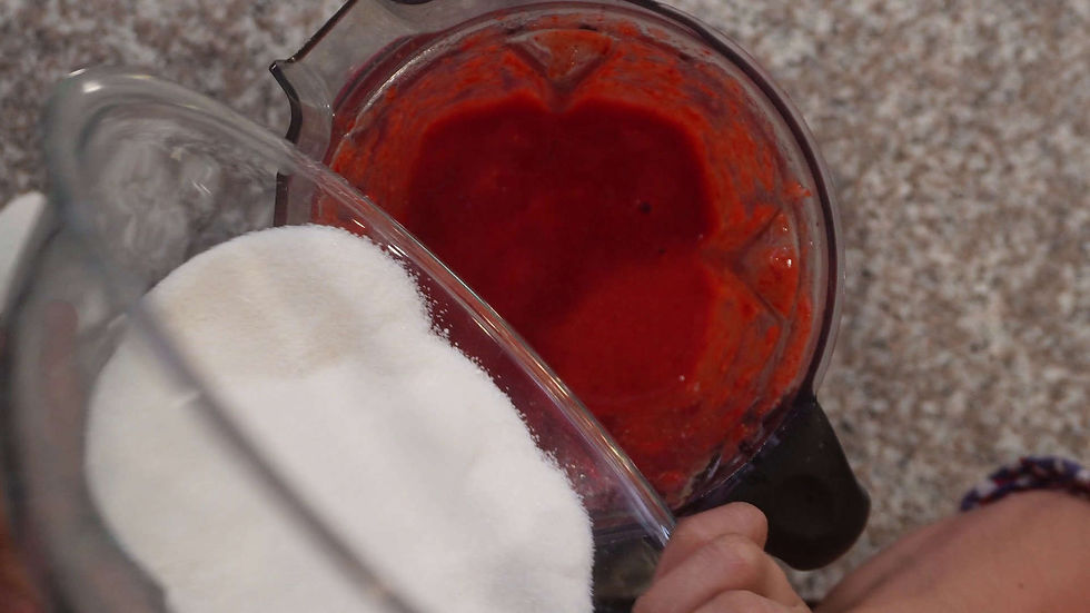 Sugar being poured into a blender with red mixture. Blurry countertop background. A hand holds the blender. Cozy kitchen scene.