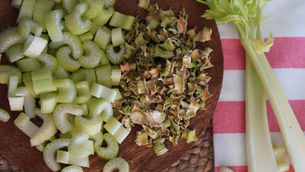 Chopped fresh and dried celery on a wooden board, with whole stalks on a red-and-white striped cloth in the background.