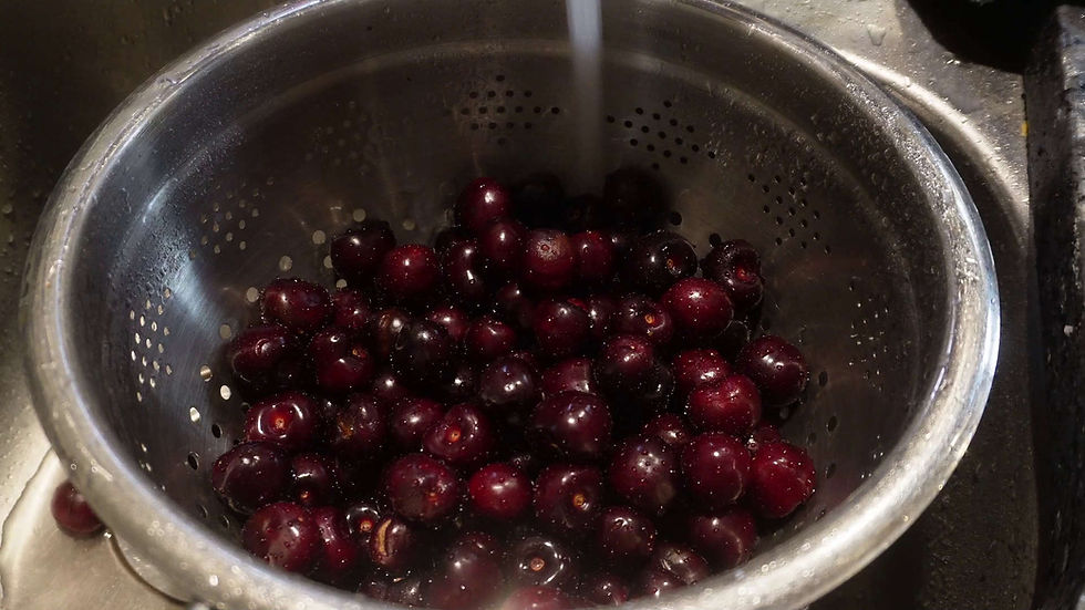 Dark cherries being rinsed in a metal colander under running water, in a kitchen sink. The cherries appear fresh and glossy.