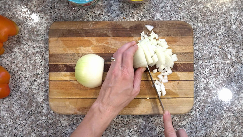 Hands chopping a white onion on a wooden cutting board. Granite countertop, diced onions, and red bell peppers visible in the background.