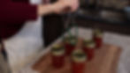 Person in a kitchen uses tongs to lift a jar of red jam with gold lids. Granite countertop, brown mat, and sink in background. Cozy mood.