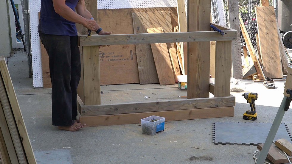 Barefoot person assembles a wooden frame outdoors, with drill, screws, and lumber around a bright DIY work area.
