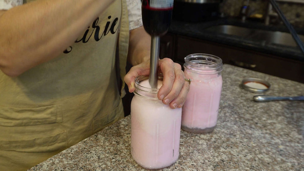 Person uses a blender to mix pink liquid in a jar on a kitchen countertop. Wearing an apron with partial text "ir" visible.