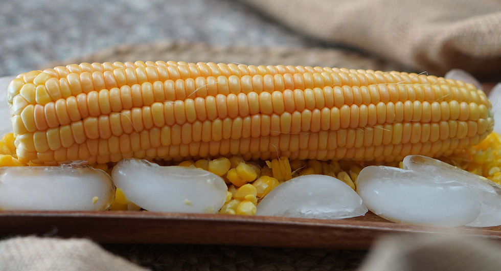 Close-up of a yellow corn cob on ice and kernels, set on a wooden tray. The background is blurred with a neutral, textured fabric.