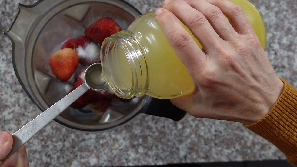 Hands pour lemon juice from a jar into a blender with strawberries. Granite countertop background, close-up of a teaspoon.