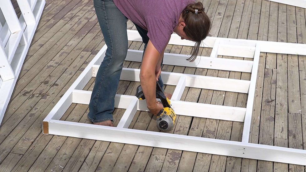 Woman in a purple shirt and jeans uses a nail gun to assemble a white wooden frame on a weathered deck.
