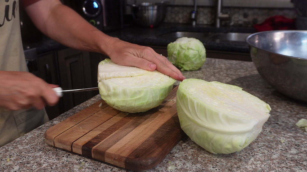 Hands slicing a cabbage on a wooden cutting board in a kitchen. A metal bowl and another cabbage in the background on a granite countertop.