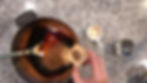 Hand adding cinnamon to dark sauce in a pot on a speckled countertop. A wooden spoon stirs. Empty cup and metal lid nearby.