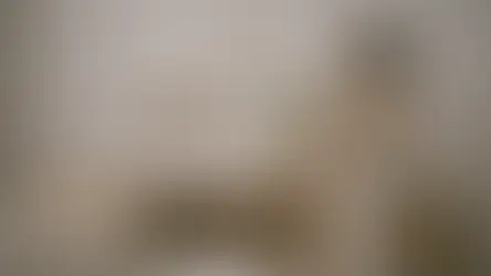 mujer colocando un jarrón en un mueble de una habitación. Hay dos cuadros en la pared, varios libros y un sofá. Todo con colores beige