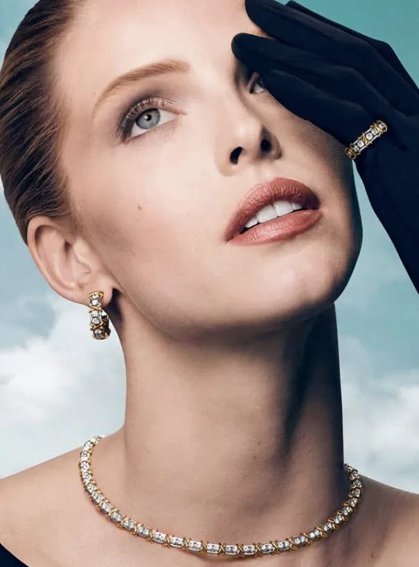 mujer con el pelo recogido y mirando hacia arriba, lleva unos guantes oscuros, unos pendientes, un collar y un anillo