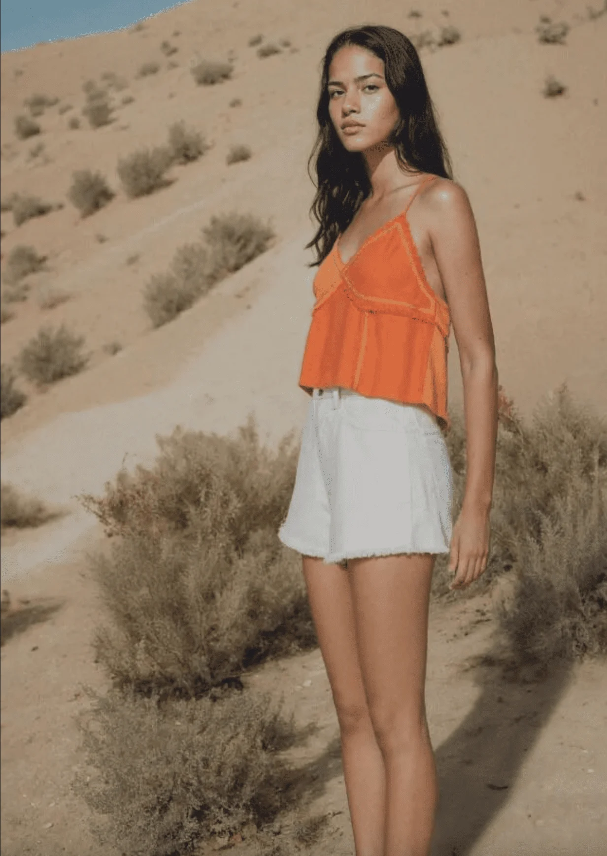 Mujer con el pelo largo moreno lleva un top naranja y un pantalón blanco. Posa en un desierto.