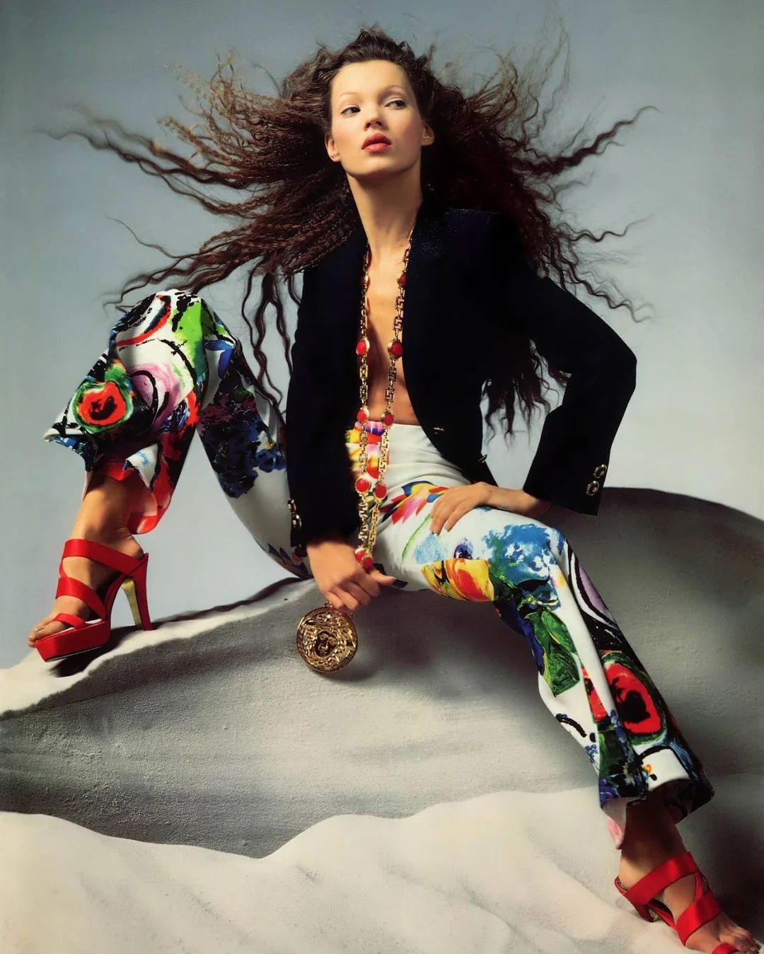 chcia con el pelo rizado y largo vestida con chaqueta negra y pantalones multicolor está sentada sobre arena