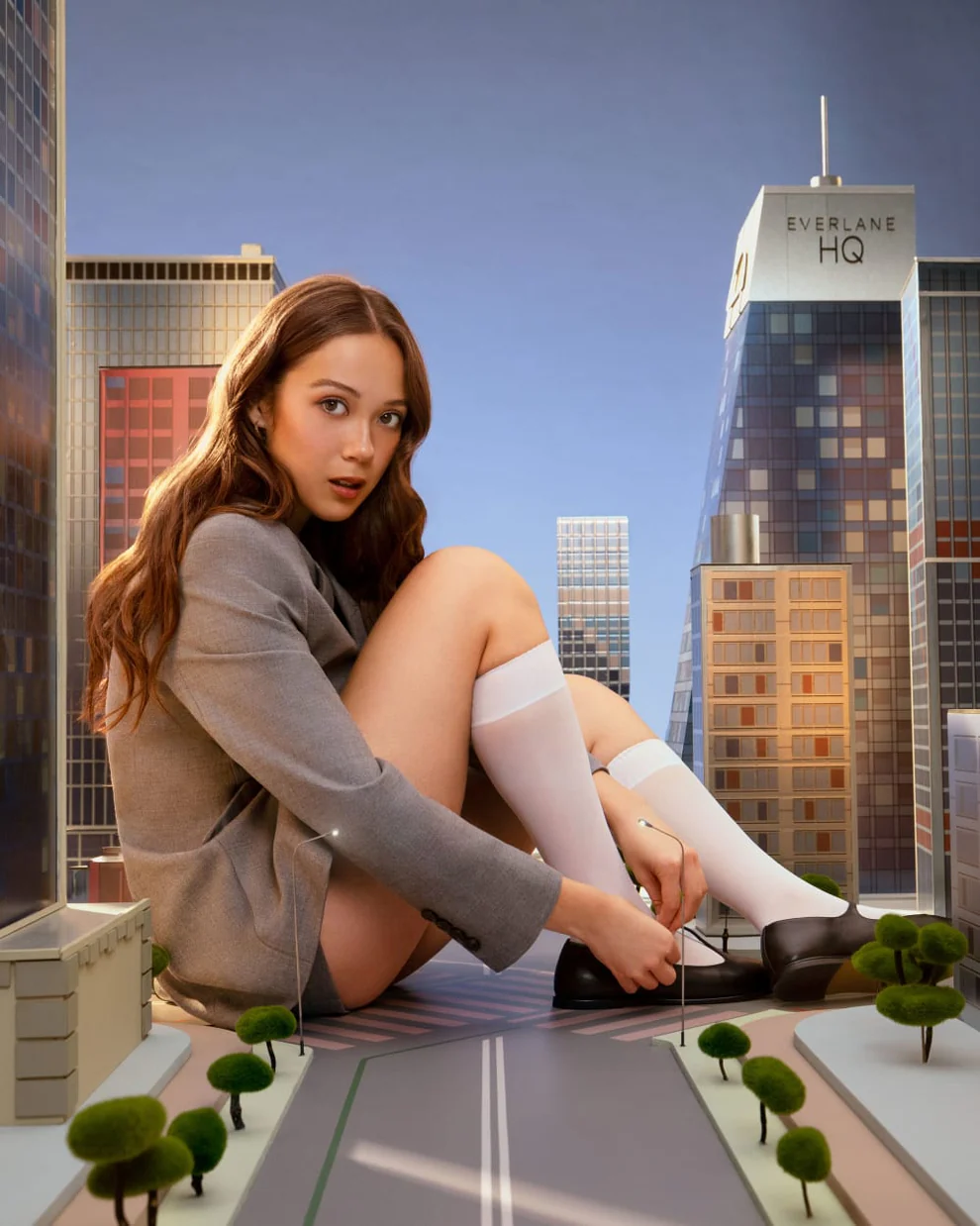 Girl sitting in a miniature city. She is wearing a gray jacket, long white socks, and black shoes.