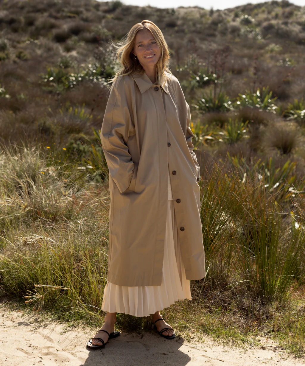 Woman outdoors wearing a long beige coat, a long white skirt, and sandals