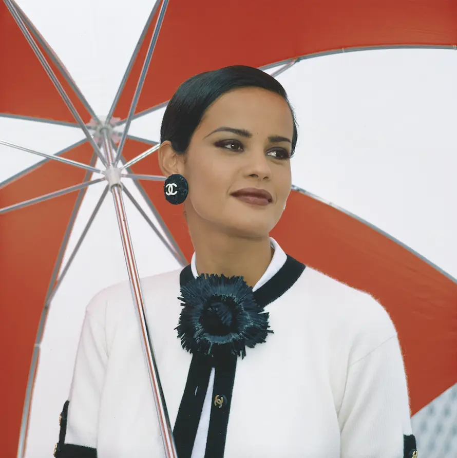 chica con el pelo recogido, pendientes negros, camisa blanca y accesorio negro lleva un paraguas blanco y rojo.