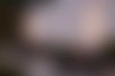 Terraza con dos mesas. Cada mesa tiene cuatro sillas, velas y una maceta pequeña. Hay varias plantas y una pared blanca donde hay dos lámparas de pared encendidas.