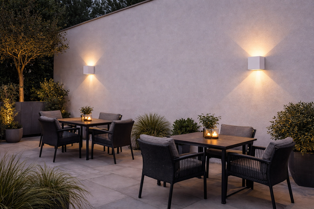 Terraza con dos mesas. Cada mesa tiene cuatro sillas, velas y una maceta pequeña. Hay varias plantas y una pared blanca donde hay dos lámparas de pared encendidas.