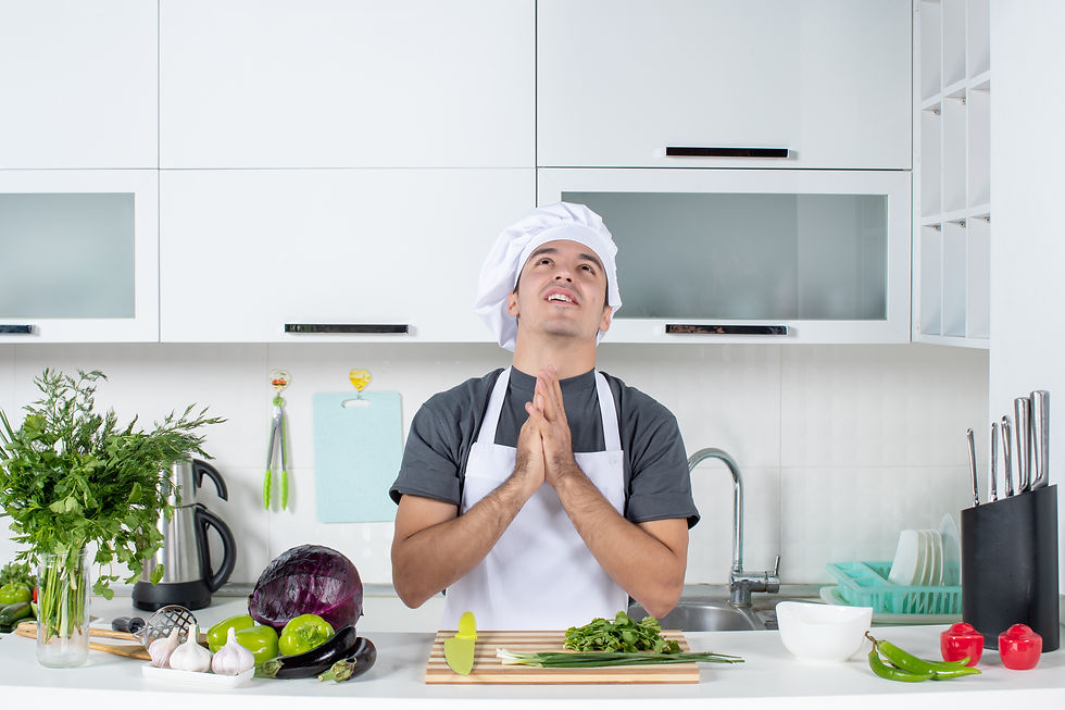 Chef in white hat and apron, looks upwards with clasped hands, surrounded by fresh vegetables in a bright kitchen. Mood is hopeful.