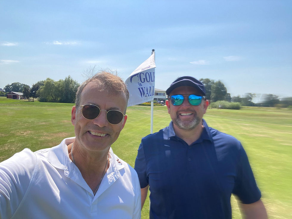 Two smiling men in sunglasses pose at a sunny golf course. A flag reads "GOLF". A clubhouse and trees are in the background.