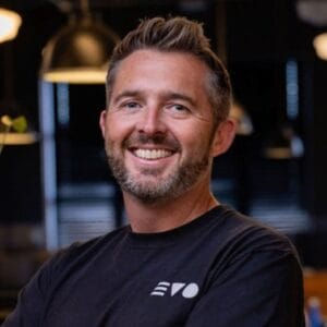 Smiling man with short hair and beard in a black EVO shirt in a dimly lit room with industrial lamps in the background.