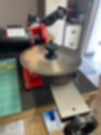Circular saw sharpening machine with a large metal blade on a workbench. Background shows storage cabinets and boxes.