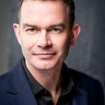 Man in a dark blazer smiles slightly against a gray textured backdrop. The mood is professional and confident. No text visible.
