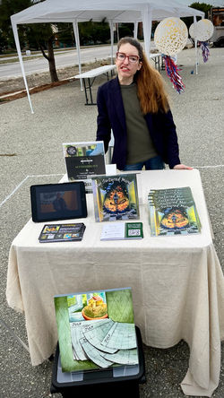 Woman (author) wearing black coat and red glasses is standing behind booth for an outdoor market, wi