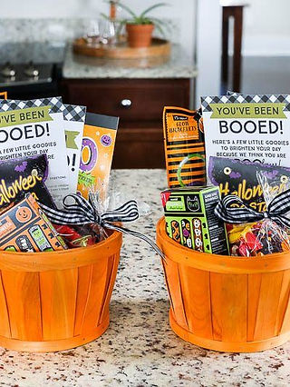Two bright orange Halloween baskets filled with festive goodies, including candy, activity packs, and “You’ve Been Booed!” signs. Each basket is decorated with a black-and-white gingham ribbon and sits on a kitchen counter, ready to be delivered as part of a fun neighborhood “boo” surprise.