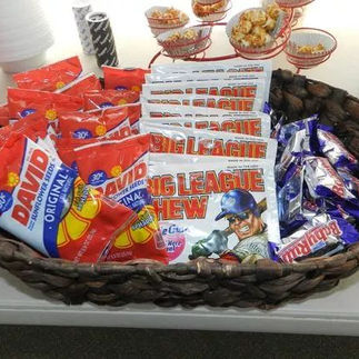 Woven basket filled with classic baseball game snacks including David sunflower seeds, Big League Chew bubble gum, and Nestlé Crunch bars. The basket is placed on a table alongside red cups and other treats, creating a fun and nostalgic snack station.