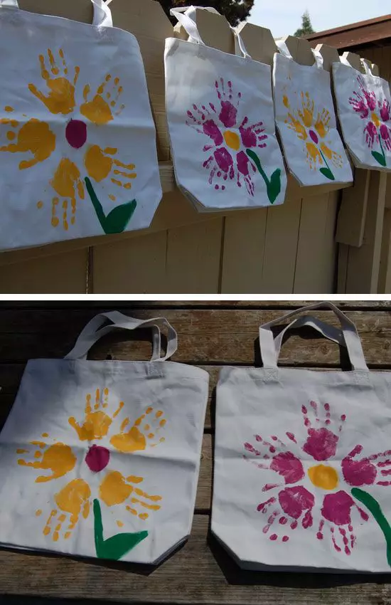 Two canvas tote bags featuring large handprint flowers in pink and yellow with green stems, shown on a wooden surface.