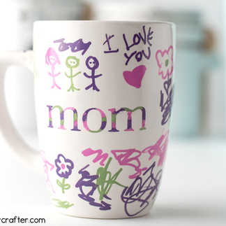 White mug decorated with colorful childlike drawings, hearts, and the words “I Love Mom,” designed as a handmade gift, using Sharpie markers.